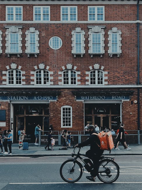 A person riding a black bicycle along a street in front of a red-brick building with multiple white-framed windows and a sign indicating Waterloo Road and train station, with pedestrians walking nearb