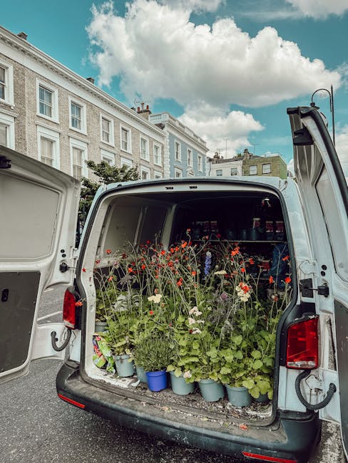 The image depicts the open back of a white delivery van filled with a variety of fresh flowers and green foliage, including tall stems of red poppies, white daisies, and lush green leaves in blue and