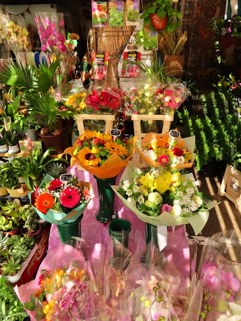 A display of fresh flower bouquets arranged on a pink table inside a florist shop, showcasing a variety of floral gifts. The arrangement includes bright yellow sunflowers with orange and pink gerbera