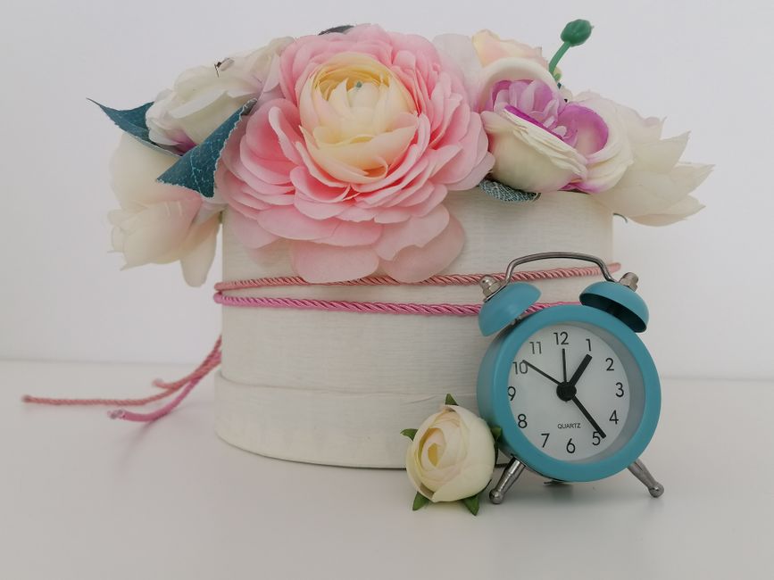A floral arrangement featuring pink and white roses, cream-colored peonies, and soft blue accents in a white round hat box with pink ribbon, placed on a white surface. Beside the box is a small white 