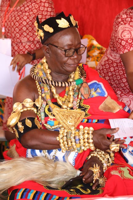 A woman dressed in traditional African attire, featuring vibrant red and black clothing with intricate patterns, adorned with numerous gold and beaded jewelry including necklaces, bracelets, and a cro