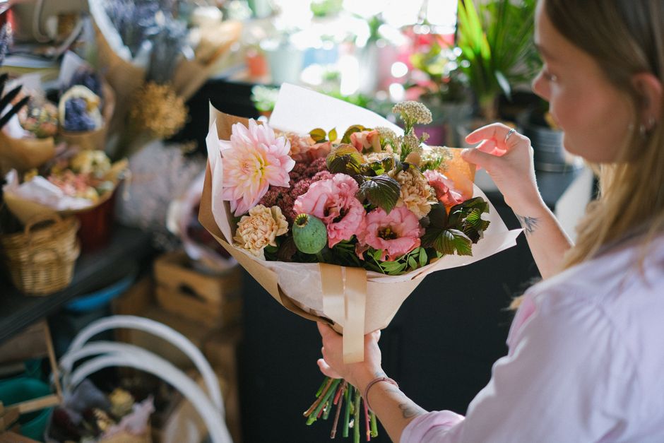 A woman with long, light brown hair wearing a light pink shirt holds a freshly arranged bouquet of flowers in a florist shop. The bouquet includes large pink dahlias, soft pink roses, cream-colored ca