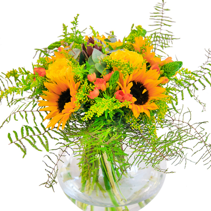 Inspired by the charm of Canonbury's neighbourhood gardens, this vibrant bouquet bursts forth with sunflowers that command attention-each bloom a beacon of golden light, framed by velvety dark centres. Clusters of bright yellow filler flowers, reminiscent of solidago or mimosa, weave through the arrangement, adding depth and highlighting the sunflowers' exuberance. Coral-orange accent blooms peer out with a gentle confidence, suggesting the creativity of the area's artists and residents. The design is given a decidedly lush feel by the abundance of rich greenery; feathery fern fronds dance over the edges, while silvery eucalyptus and lively lime foliage provide a crisp, modern note. Nestled in a clear, round glass vase, the bouquet's stems are visible in their full, fresh glory-much like the inviting transparency seen in Canonbury's friendly cafés. Evoking sunshine and comfort, the bouquet's subtle fragrance stirs up memories of community picnics in Highbury Fields. Delivered with care by local experts, this arrangement is perfect for birthdays, anniversaries, or spontaneous displays of affection, carrying a bit of Canonbury's sunshine and community spirit into any space.